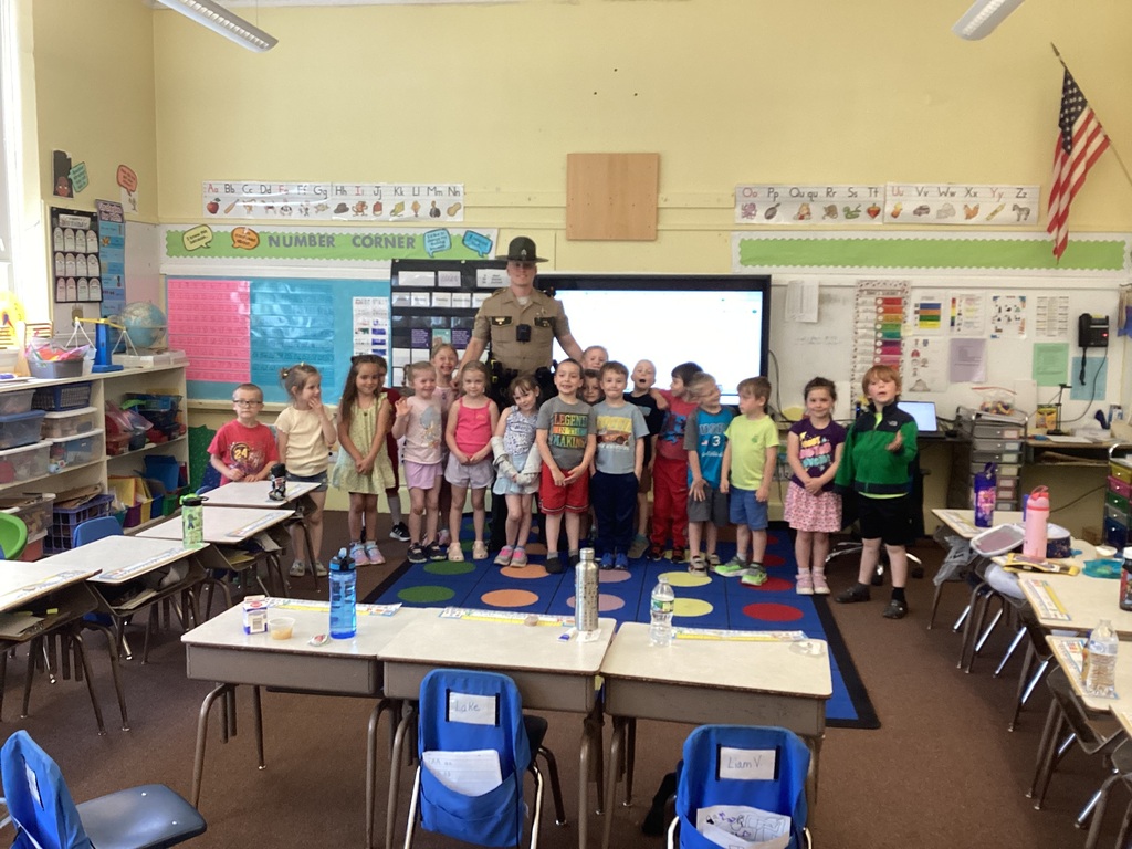 A Vermont State Trooper stands with a kindergarten class inside a Fair Haven Grade School classroom. Students smile and gather around the officer on a colorful classroom rug while posing for a group photo.
