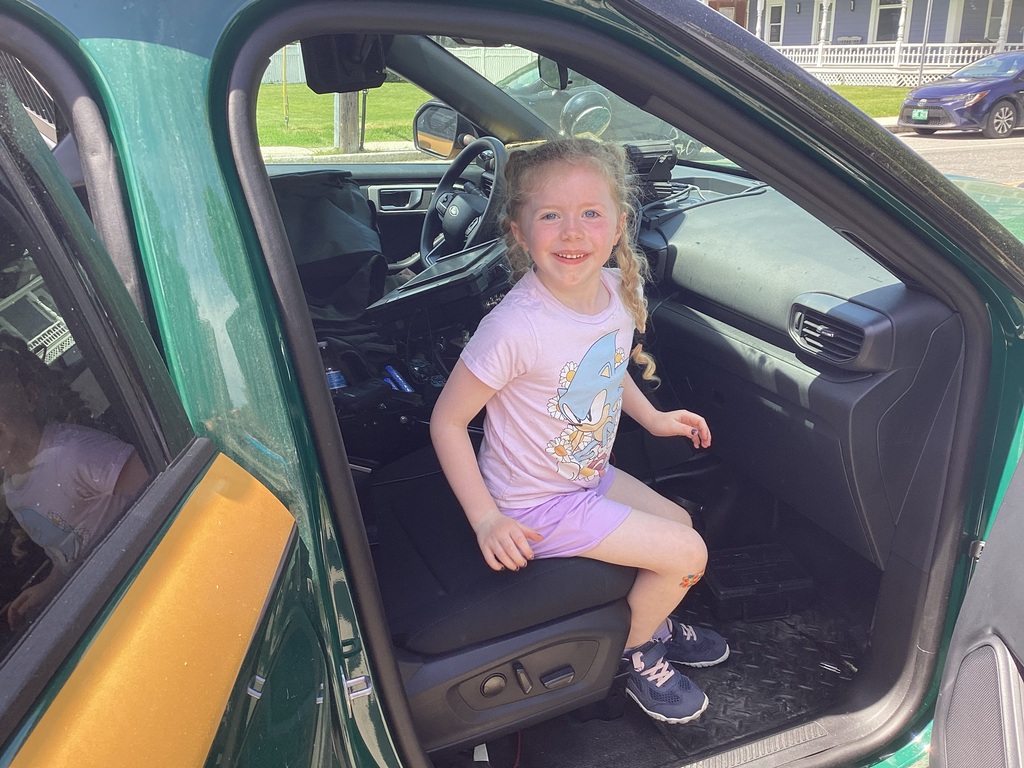A kindergarten student smiles while sitting inside the passenger seat of a Vermont State Police cruiser during a classroom visit. The vehicle's interior and dashboard are visible.