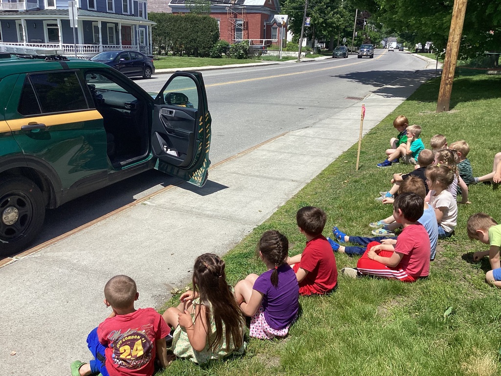 A group of kindergarten students sits on the grass near a sidewalk watching a demonstration involving a Vermont State Police cruiser parked nearby with its door open. The children observe as they learn about the police vehicle and its equipment.