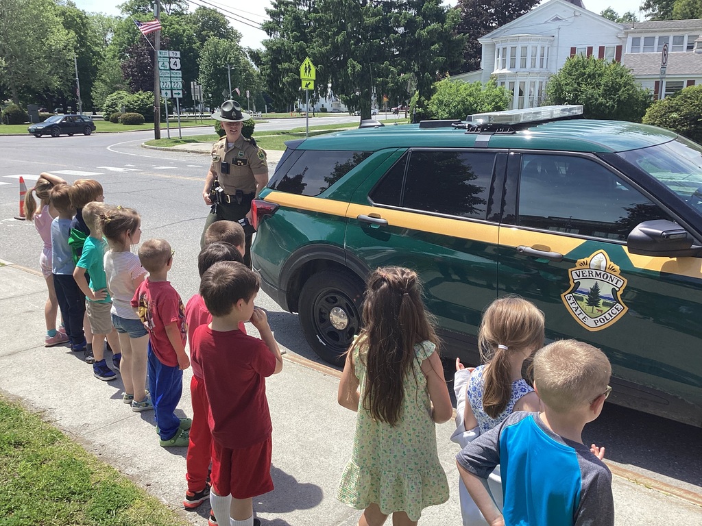 Kindergarten students stand on a sidewalk outside as a Vermont State Trooper explains features of a Vermont State Police cruiser. The children listen attentively while gathered around the vehicle on a sunny day.