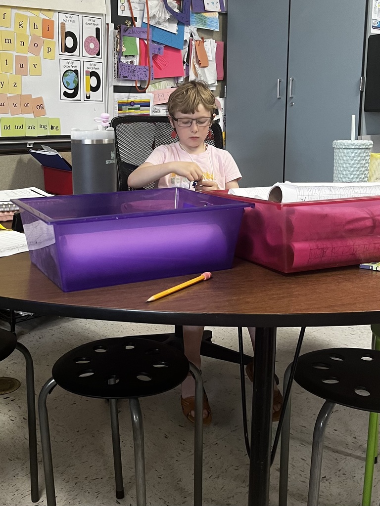 A child wearing glasses, red earrings, and a pink shirt sits focused at a round wooden classroom table, working on a small craft or object with their hands. In the foreground on the table, there is a yellow pencil, a large translucent purple plastic bin, and a translucent pink plastic bin containing rolled-up papers. The background features a classroom whiteboard covered in educational alphabet cutouts and posters.