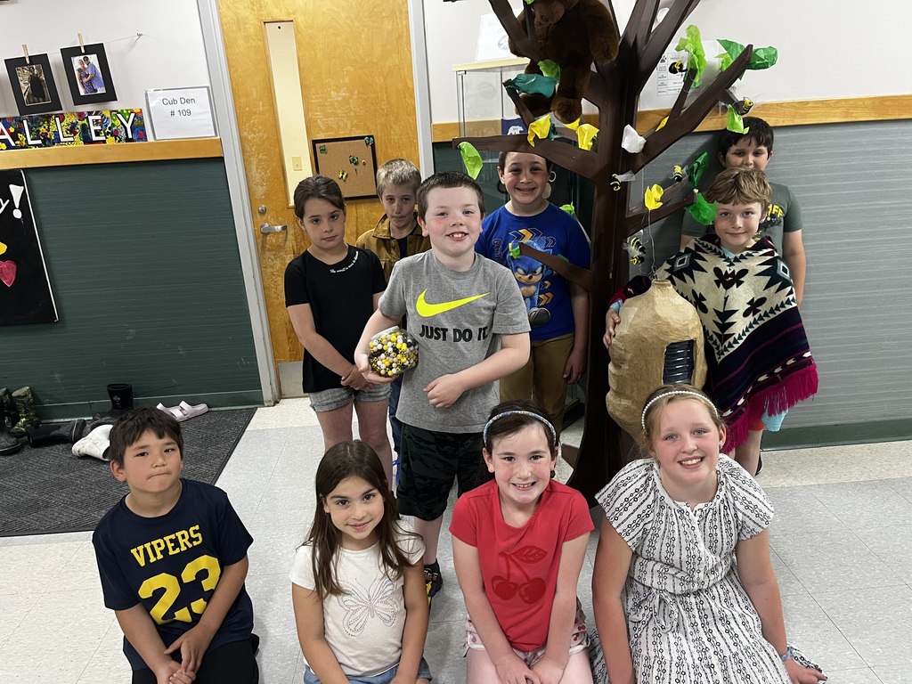 A group of nine elementary school children pose together for a photo in a classroom or school hallway. They are gathered around a decorative cardboard tree that is adorned with paper bees and features a large, handmade paper beehive hanging from a branch. One boy in a grey Nike shirt stands near the center holding a clear jar filled with yellow and black marbles or beads, while the other children smile or pose from seated and standing positions around him.  
