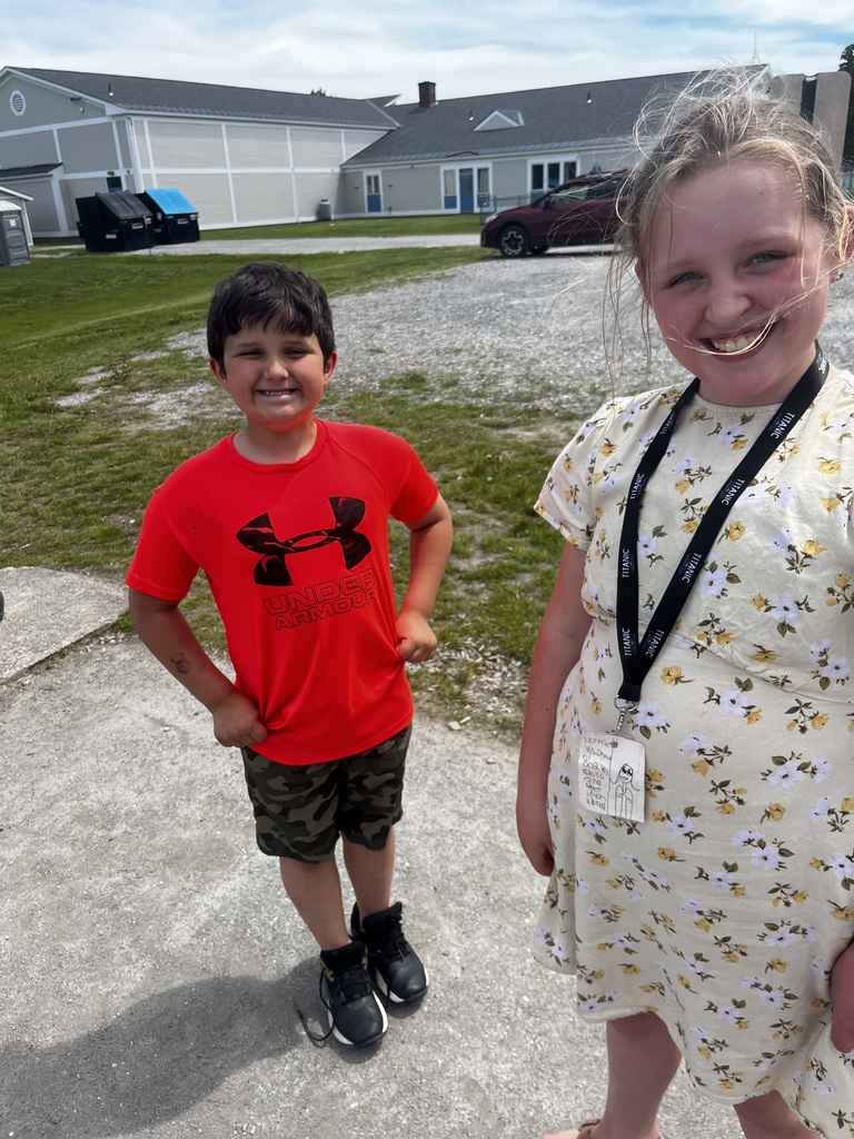 Two smiling children stand outside on a gravel driveway in front of a light grey school building with blue doors. On the left, a boy in a bright orange Under Armour shirt and camouflage shorts poses with his hands on his hips. On the right, a girl wearing a yellow floral dress and a black lanyard smiles widely at the camera, with the wind blowing a few strands of hair across her face. 