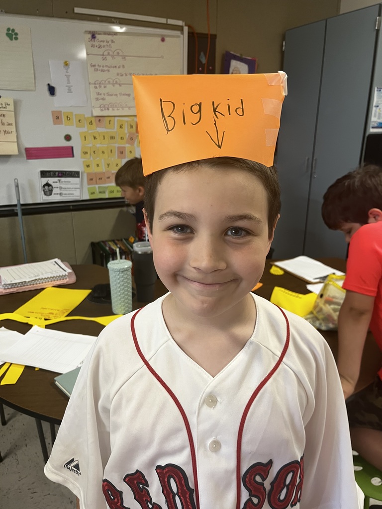  A young boy with short brown hair smiles warmly at the camera inside a classroom. He is wearing a white Boston Red Sox baseball jersey with red trim. Taped to the top of his head is a bright orange piece of paper folded into a crown shape, featuring the words "Big kid" handwritten in black marker with an arrow pointing downward toward his face. 