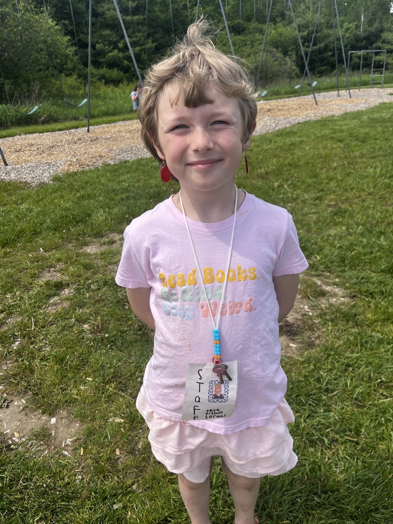  A medium shot of a young child with short blonde hair smiling while standing outdoors in a grassy area with a playground structure visible in the background. The child is wearing a pink T-shirt that reads "Read Books. Be Kind. Stay Weird." in colorful lettering, light pink shorts, red dangling earrings, and a handmade "STAFF" ID badge on a beaded white lanyard around their neck.  