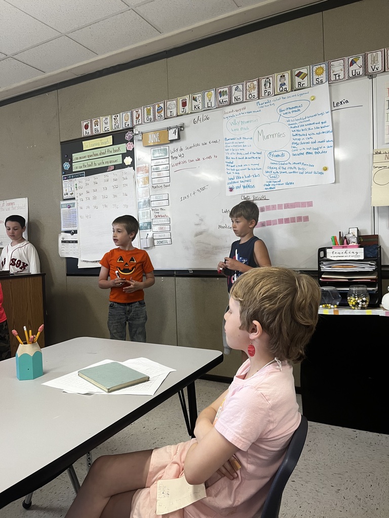 An indoor shot of a classroom presentation or activity. In the foreground, a child with short blonde hair and red earrings sits at a white table with their arms crossed, looking toward the front of the room. In the background, two young boys stand near a large whiteboard filled with diagrams and handwritten notes about "Mummies" and "Pyramids," while another boy in a baseball jersey stands off to the far left.