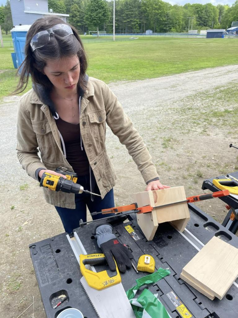 A student wearing safety glasses uses a drill to secure pieces of a birdhouse that are clamped together on a portable workbench.