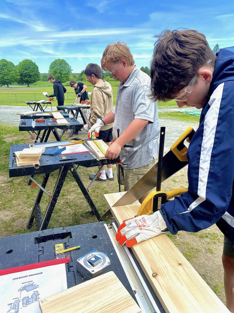 Several students work at individual stations outdoors, measuring, cutting, and assembling wood as part of a birdhouse engineering challenge.