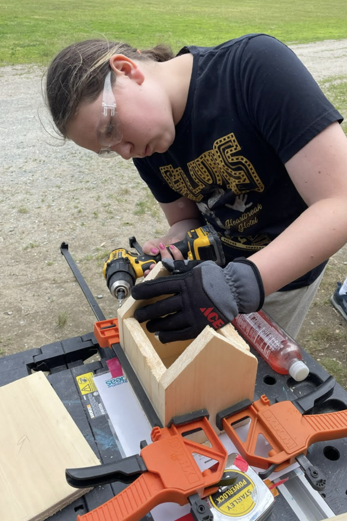 A student uses a cordless drill to assemble a wooden birdhouse at an outdoor workstation while participating in a hands-on engineering project.