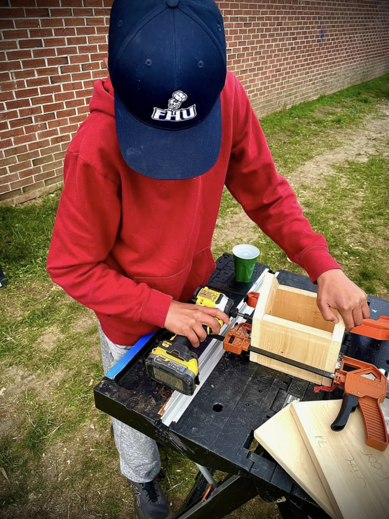 A student in a red sweatshirt and baseball cap uses a cordless drill while assembling a wooden birdhouse at an outdoor workstation.