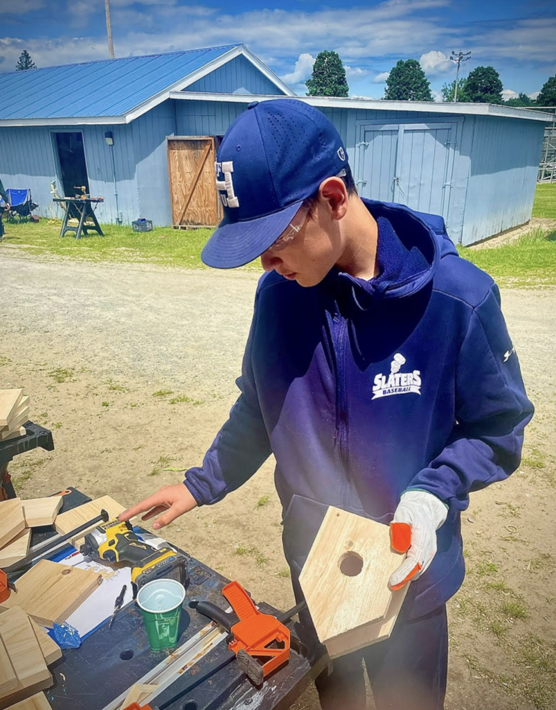 A student wearing safety glasses and gloves works on assembling a birdhouse, holding a wooden panel while standing at a workbench outdoors.
