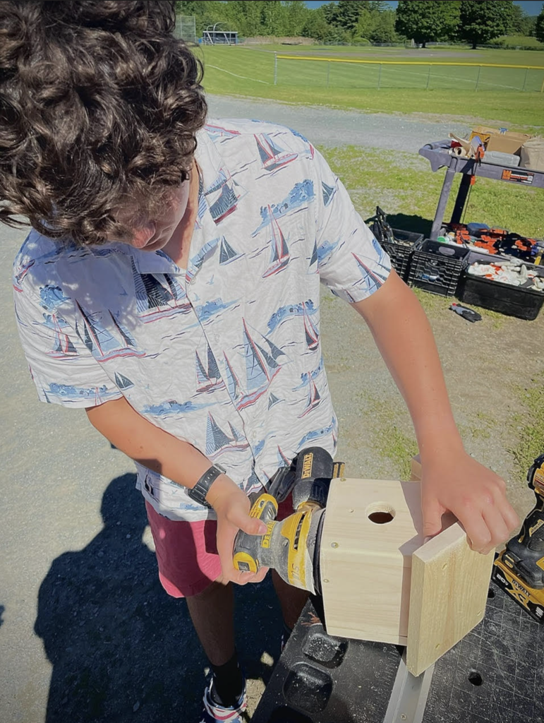 A student uses a cordless sander to sand a wooden birdhouse at an outdoor workstation while participating in a hands-on engineering project.