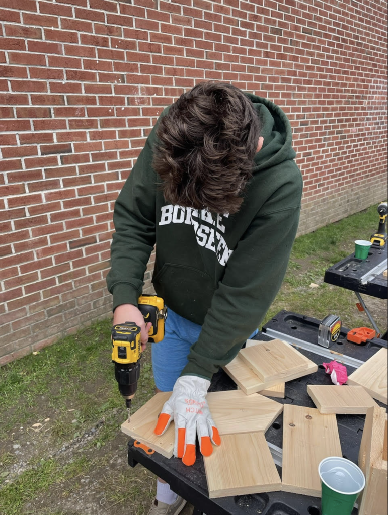A student wearing gloves uses a cordless drill to assemble wooden birdhouse components at a workstation set up outside near a brick school building.