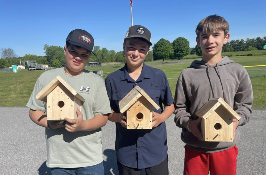Three students pose outdoors holding finished birdhouses. Athletic fields and trees can be seen in the background on a sunny day.