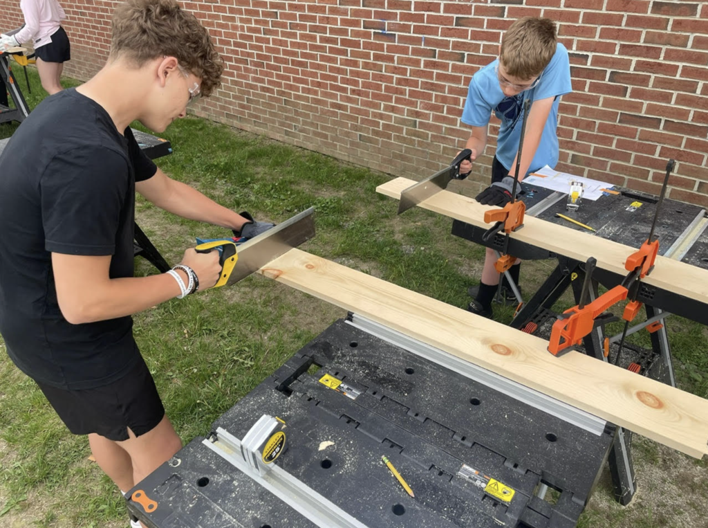 Two students use hand saws to cut wooden boards at separate workstations while constructing birdhouses during a Design Lab project.
