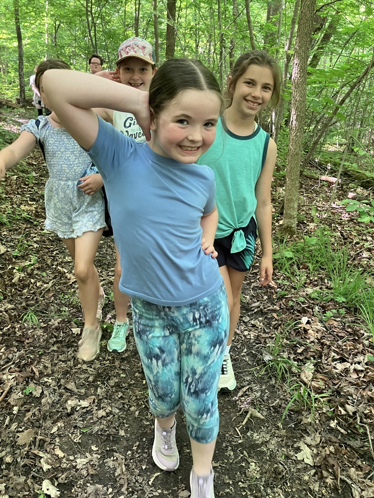 A young girl with brown hair wearing a light blue shirt and blue pants poses while hiking. There is a group of students behind her all smiiling