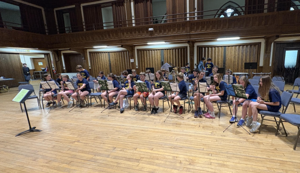 Orwell Village School band students sit in rows inside a large historic hall during a rehearsal or performance setup. Students hold woodwind and brass instruments while music stands are arranged in front of them on a wooden stage floor.