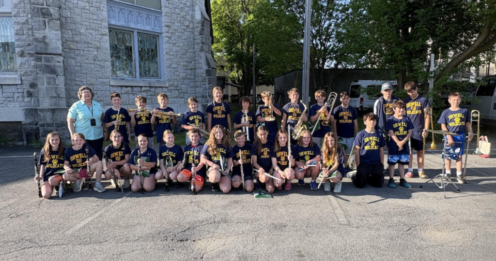 A group of Orwell Village School band students and their music teacher pose outside a stone church before a performance. Students wear matching Orwell Wildcats shirts and hold a variety of instruments, including clarinets, flutes, trumpets, saxophones, and trombones.