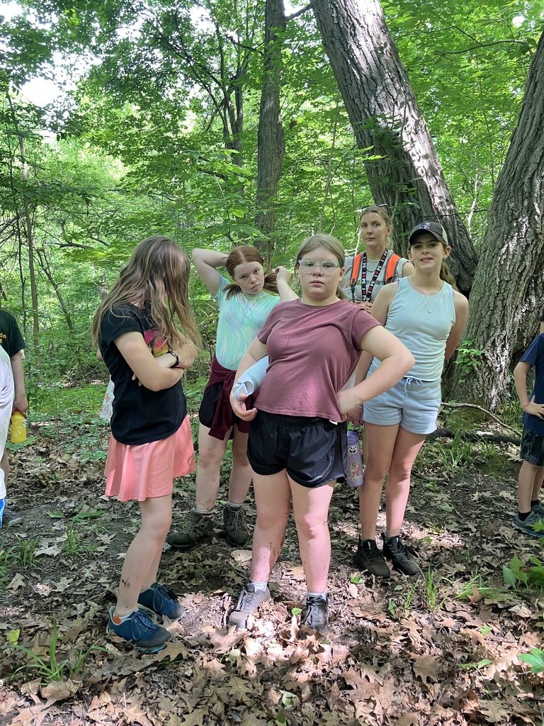  A group of five young girls and an adult woman standing together on a shaded, leaf-covered dirt trail in a dense forest. The girls are dressed in summer clothes like t-shirts, shorts, and skirts, with one girl in the center posing with her hands on her hips. Large mature trees and lush green foliage surround them.  