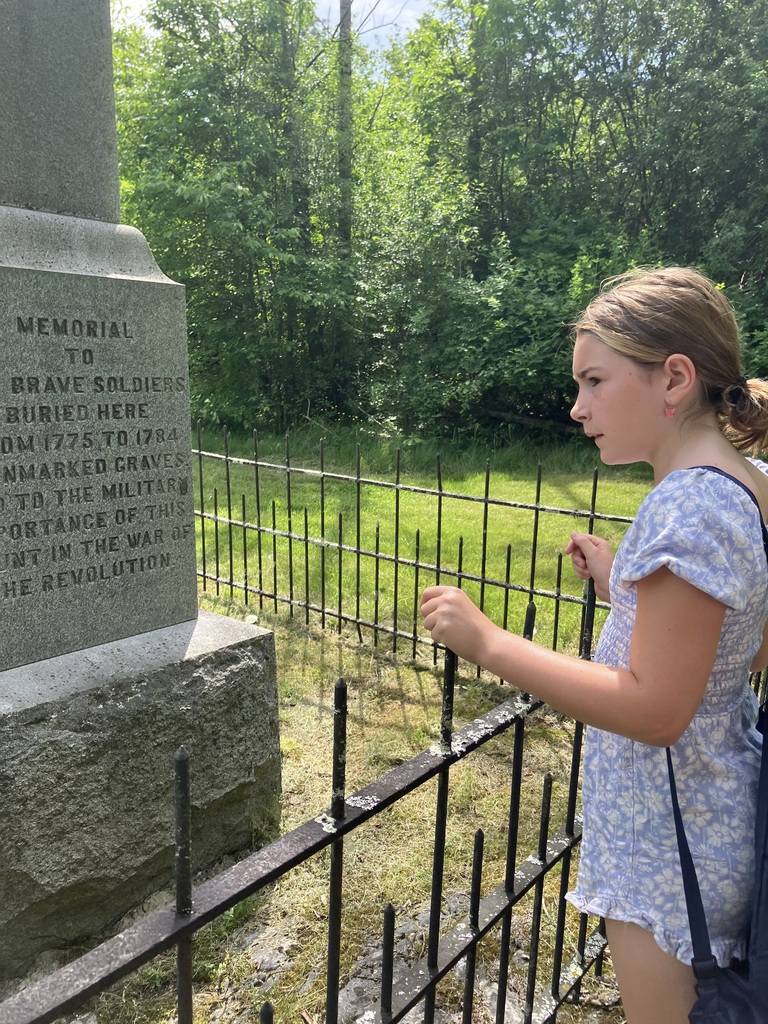 A close-up profile view of a young girl with her hair tied back, wearing a blue floral romper. She is standing next to a black wrought-iron fence, intently reading the inscription carved into a large, historic stone monument. Green woods serve as the background. 