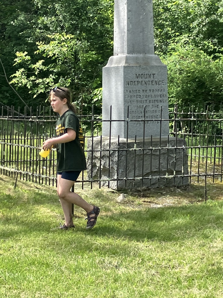 A young girl wearing a dark green t-shirt and navy shorts walks across a bright green lawn, holding a yellow cup. In the background stands a tall, historic grey stone obelisk monument enclosed by a black wrought-iron fence, bordered by dense green trees.  IMG_3389.jpg Alt Text: A group of five young boys standing together on a forest path covered in fallen leaves and soil. The boys are dressed in t-shirts, shorts, and baseball caps, with some looking directly at the camera. Tall, thick tree trunks and vibrant green foliage frame the scene.
