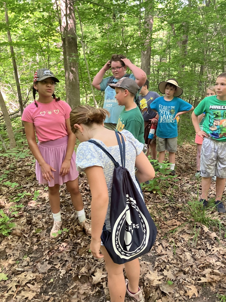 A group of children gathered on a wooded hiking trail covered in fallen leaves. In the foreground, a girl with her back to the camera wears a blue and white patterned top and a dark drawstring backpack. Other children stand around talking, wearing casual summer clothes and hats, surrounded by bright green forest trees.  