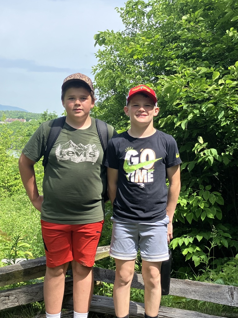  Two boys smiling and sitting on an old wooden split-rail fence outdoors. The boy on the left wears a wide-brimmed sun hat, a bright blue t-shirt, and grey shorts. The boy on the right wears a camo baseball cap, a grey "Coyotes" t-shirt, dark jeans, and holds a blue water bottle. A lush green forest and distant hills are visible under a bright sky in the background. 