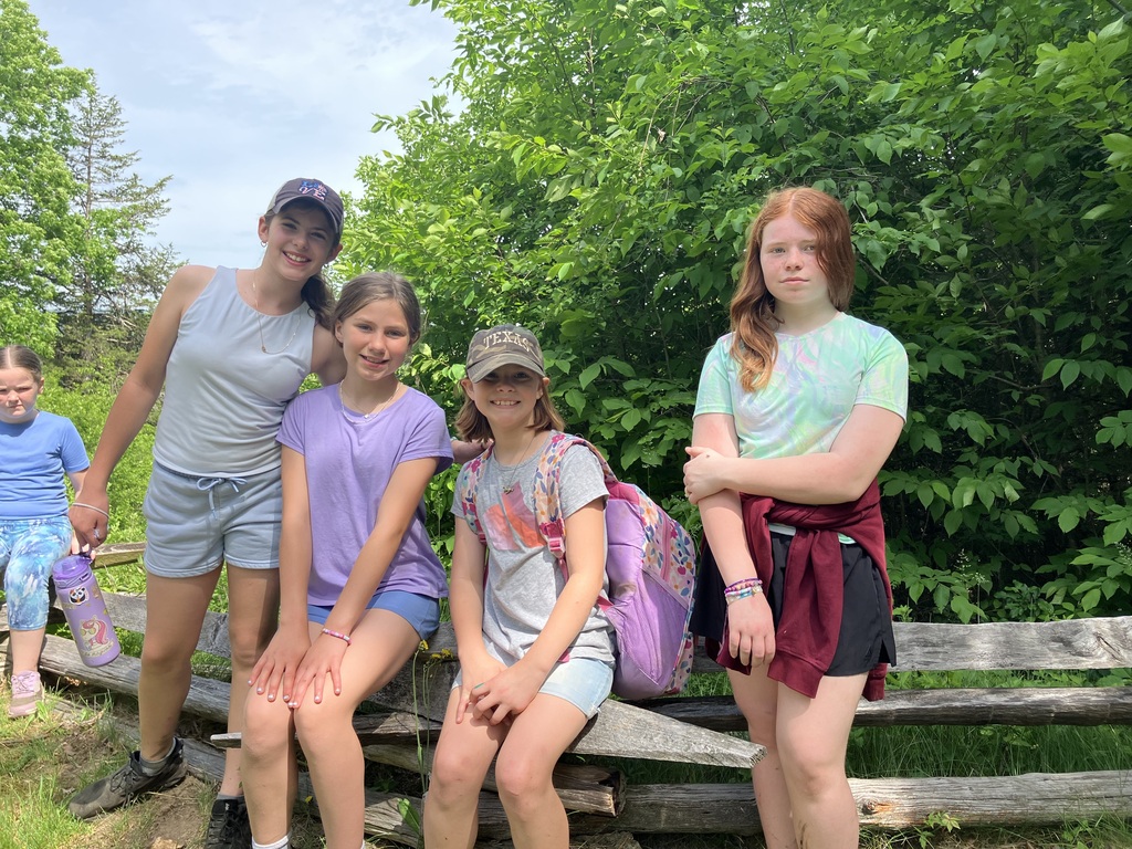  Four girls posing together against a rustic wooden split-rail fence with dense green bushes behind them. From left to right: a girl in a white tank top and grey baseball cap smiles; a girl in a purple t-shirt sits on the fence; a girl in a grey t-shirt, camo hat, and floral backpack smiles; and a girl with red hair stands with crossed arms wearing a light green patterned t-shirt with a maroon sweatshirt tied around her waist. A fifth girl in a blue shirt is partially visible on the far left.  
