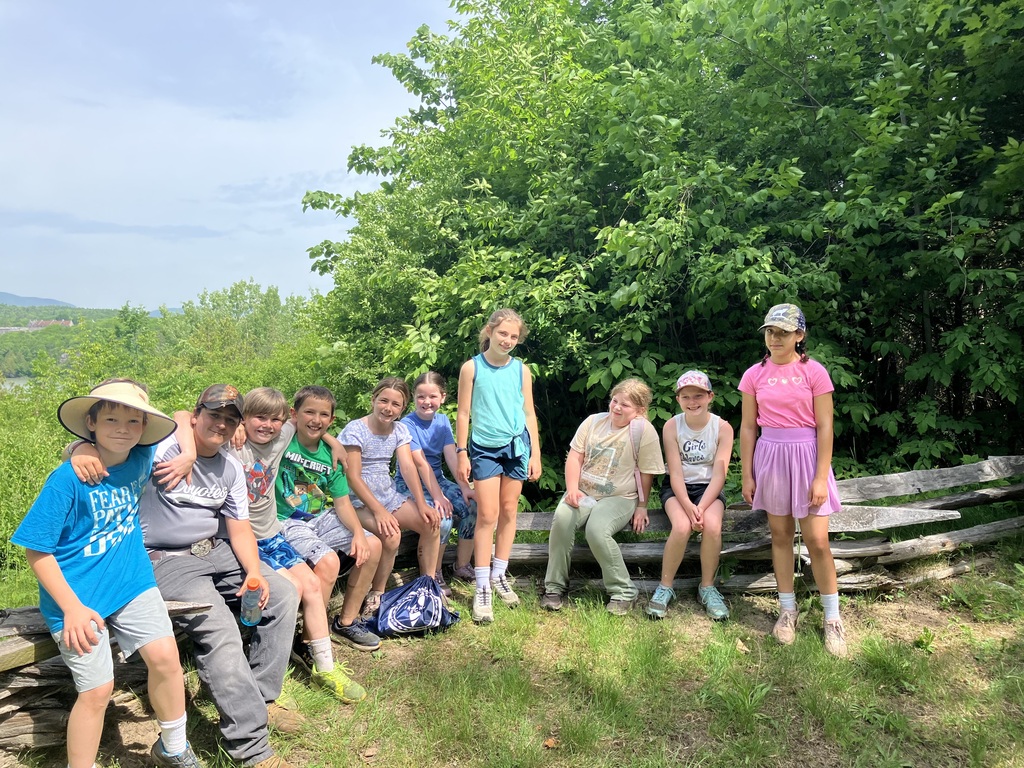 A group of ten children sitting and standing along an old wooden split-rail fence in a grassy area. They are dressed in casual summer clothes and hats, smiling for the camera. Behind them is a thick wall of green trees and bushes, with a scenic view of distant hills appearing on the far left under a slightly overcast sky. 