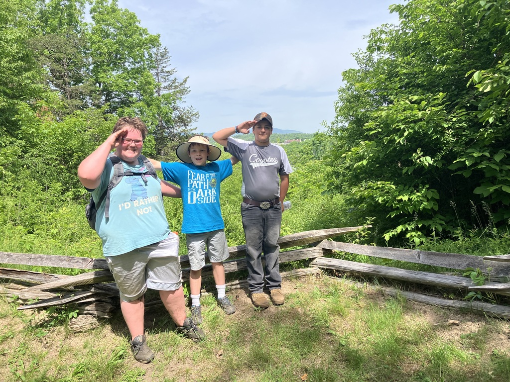  Three boys posing playfully by a wooden split-rail fence in a grassy, wooded area. The boy on the left wears a light blue t-shirt and salutes. The boy in the middle wears a sun hat and a bright blue shirt that says "Fear is the path to the dark side" with his arms outstretched. The boy on the right wears a grey "Coyotes" t-shirt and dark pants, also giving a salute.  