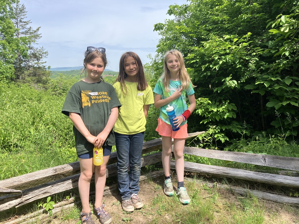 : Three girls standing together next to a wooden split-rail fence with thick green foliage behind them. The girl on the left wears a dark green "Westside Pride" t-shirt and holds a yellow water bottle. The middle girl wears a bright yellow t-shirt and jeans. The girl on the right wears a tie-dye shirt, orange shorts, and holds a blue water bottle. 