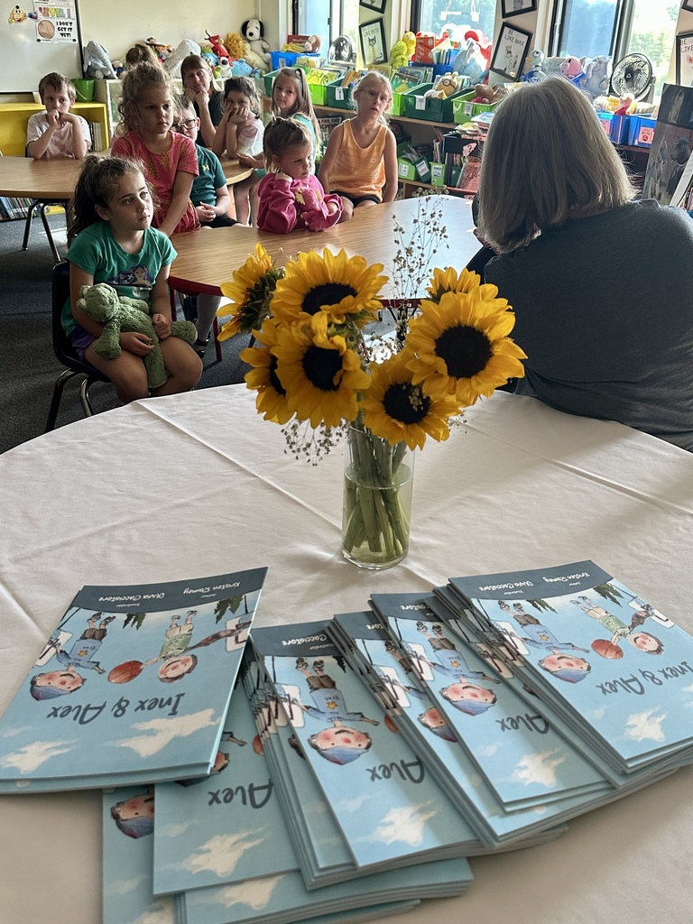 A vase of sunflowers and stacks of Inex & Alex books sit on a table as students listen to Mrs. Ramey read in the background.