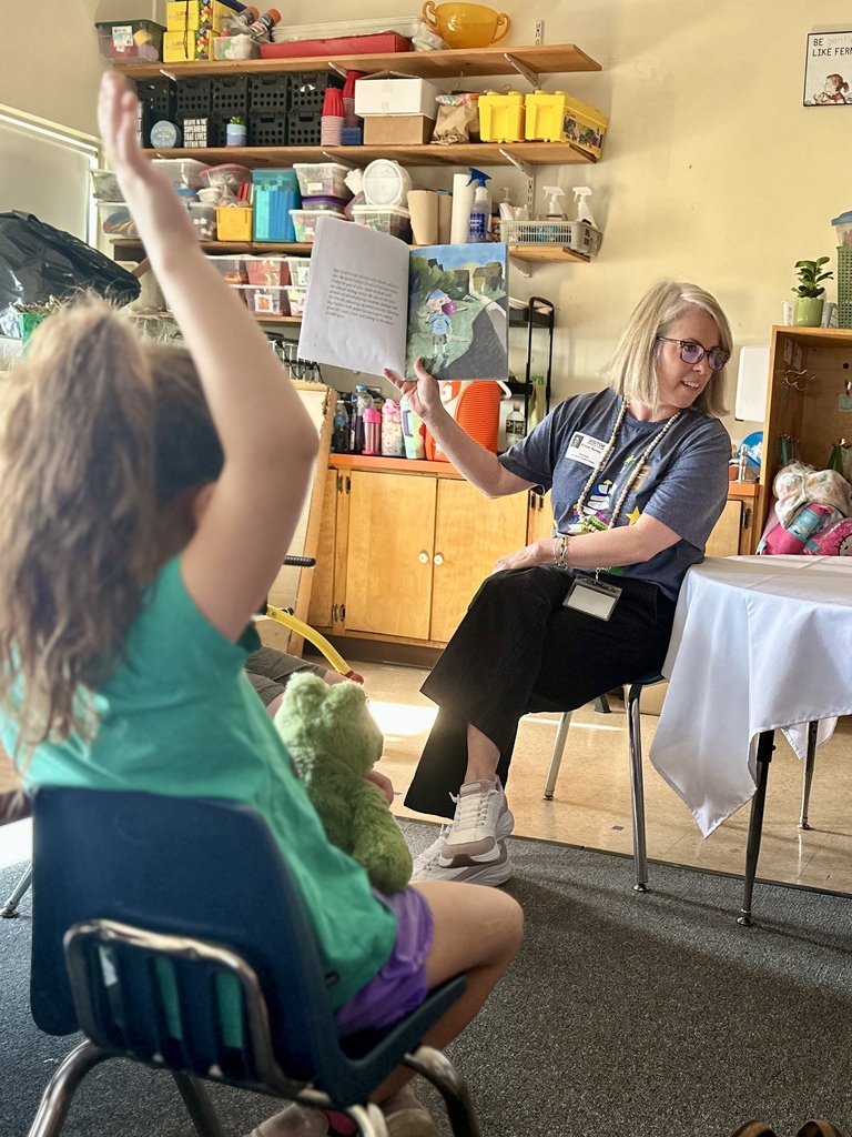 Mrs. Kristen Ramey holds up an illustrated page from Inex & Alex while reading to students. A child in the foreground raises a hand to participate.