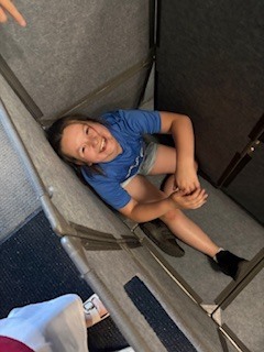 A top-down view looking into a small, enclosed space formed by gray, upholstered modular partition panels. Inside the cozy nook, a young child wearing a blue t-shirt and grey shorts sits on the floor, looking up at the camera with a wide smile.