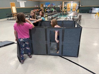  A group of elementary-aged children playing inside a large room with modular, gray felt privacy screens. Several children are gathered inside an enclosed fort-like structure made from the panels, while one boy in the foreground stands on loose panels on the floor, holding up a vertical panel to add to the structure.  