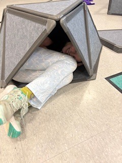  A high-angle shot of a person curled up and smiling from inside a small, triangular fort built from gray, interlocking acoustic or foam panels on a tiled floor. Only their legs in patterned white and blue leggings, light-colored sneakers with teal accents, and a glimpsed smile are visible. 