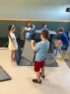 A group of elementary-aged children playing inside a large room with modular, gray felt privacy screens. Several children are gathered inside an enclosed fort-like structure made from the panels, while one boy in the foreground stands on loose panels on the floor, holding up a vertical panel to add to the structure.