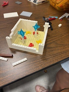 A close-up of a partially built white LEGO structure on a wooden table, resembling an aquarium or pool. Inside, translucent blue LEGO dolphins are mounted on white pillars over a tan baseplate decorated with small red and yellow coral pieces. Stray LEGO bricks scatter the surrounding tabletop. 