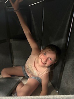 A high-angle shot of a person curled up and smiling from inside a small, triangular fort built from gray, interlocking acoustic or foam panels on a tiled floor. Only their legs in patterned white and blue leggings, light-colored sneakers with teal accents, and a glimpsed smile are visible.
