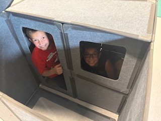 : A high-angle view looking down into an enclosed fort built from grey, fabric-covered modular panels. Two young boys are inside the structure, smiling up at the camera; one boy in a red shirt peeks through a doorway gap, while the other boy with glasses looks out from a square cutout window.ggings, holding a small plastic piece in her hands as she builds.