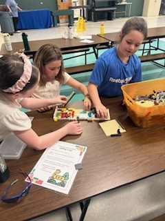  Three young girls sit closely together at a long wooden cafeteria table, collaboratively building a colorful structure out of toy bricks. A large orange plastic bin filled with building pieces sits to their right, and an instructional paper with a cartoon zoo drawing lies on the table in front of them.