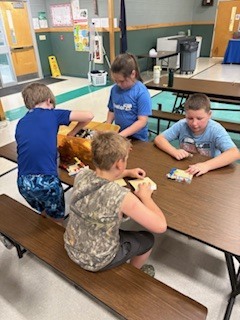  Four children sit or stand around a long wooden table in a school cafeteria or community room, working with building blocks. One boy in a blue shirt reaches into a large orange storage tub, while two other boys and a girl in a blue shirt assemble flat baseplates with various colorful pieces. 