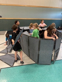 A high-angle view looking down into an enclosed fort built from grey, fabric-covered modular panels. Two young boys are inside the structure, smiling up at the camera; one boy in a red shirt peeks through a doorway gap, while the other boy with glasses looks out from a square cutout window.  
