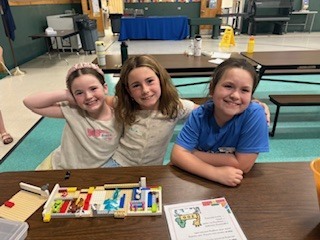 Three young girls sit closely together at a long wooden cafeteria table, collaboratively building a colorful structure out of toy bricks. A large orange plastic bin filled with building pieces sits to their right, and an instructional paper with a cartoon zoo drawing lies on the table in front of them. 