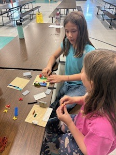  Two young girls sit at a long wooden cafeteria table, focused on building with toy bricks. The girl in the background wears a light blue t-shirt and works on a small baseplate, while the girl in the foreground wears a pink shirt and patterned leggings, holding a small plastic piece in her hands as she builds.