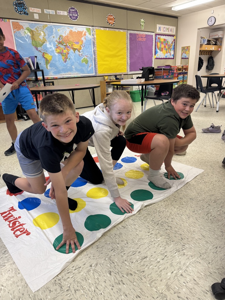 In the foreground of a classroom, three children are smiling at the camera while playing Twister. One boy in a black t-shirt and grey shorts has his hand on a green circle. Next to him, a girl in a grey hoodie has her hand on a different green circle. A second boy in an olive green shirt and red shorts crouches on the mat. The background features a classroom setting with a world map, colorful bulletin boards, and school desks.