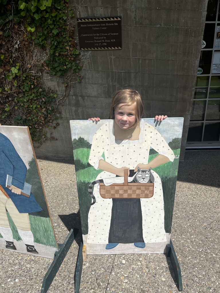  A young girl with light brown hair peeks over a painted photo cutout board outside a concrete building. The cutout depicts a woman in a colonial-style white dress with black polka dots, holding a sleeping tabby cat in a woven basket. The girl rests her hands on top of the board, looking directly at the camera with a slight smile. A bronze historical plaque is visible on the wall behind her.  IMG_6033.jpg Two young girls pose behind side-by-side colonial photo cutout boards outside a building with ivy growing on the wall. The girl on the left peeks through the cutout of a man wearing a blue coat, orange vest, and tan breeches. The girl on the right smiles from behind the cutout of a woman in a white patterned dress holding a basket with a sleeping cat. 
