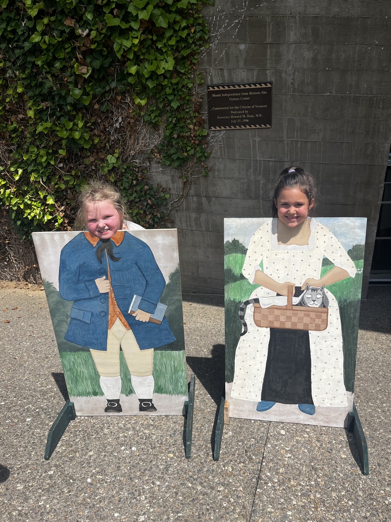 A young girl smiles while posing behind a cutout board of a colonial woman holding a basket with a cat. Behind her, two other girls stand waiting, and an empty cutout board of a man in a blue coat is visible to the left. A bronze plaque mounted on a concrete wall is visible in the background.