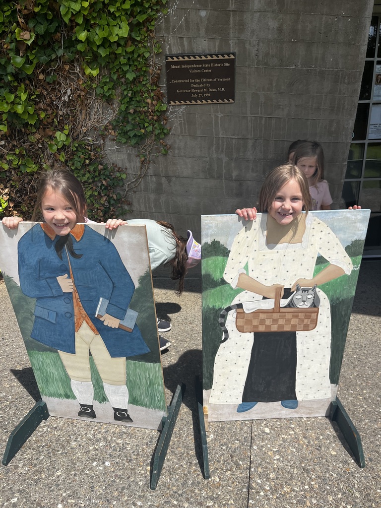 Two young girls pose behind side-by-side colonial photo cutout boards outside a building with ivy growing on the wall. The girl on the left peeks through the cutout of a man wearing a blue coat, orange vest, and tan breeches. The girl on the right smiles from behind the cutout of a woman in a white patterned dress holding a basket with a sleeping cat. 