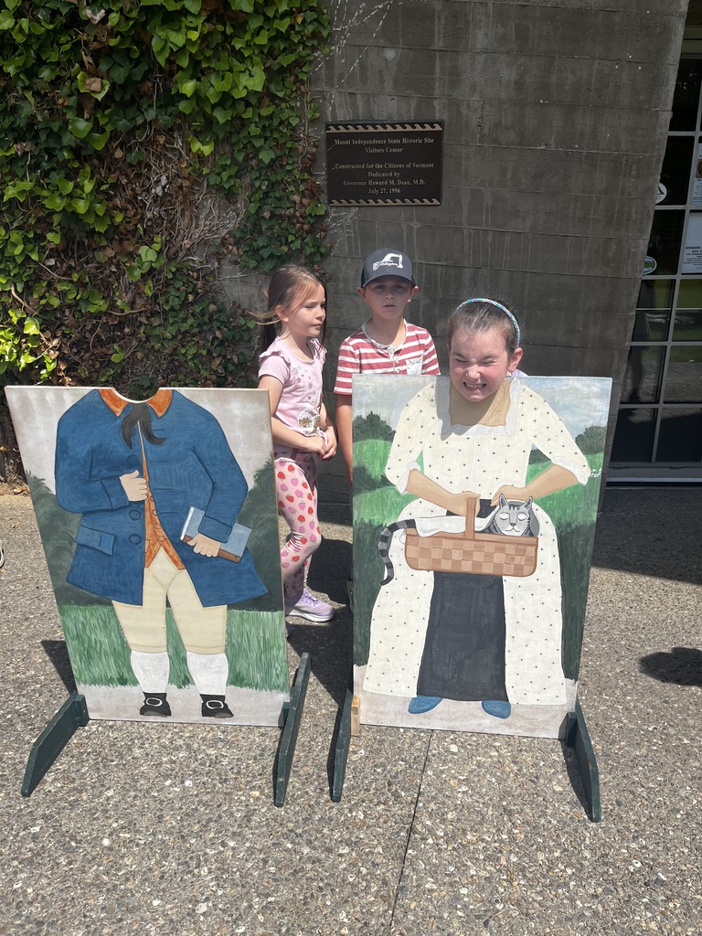  A group of children playing around two colonial photo cutout boards. Two girls smile directly at the camera while posing in the head cutouts—one as the man in the blue coat and the other as the woman holding a cat basket. In the background, another child bends over between the boards, and a fourth child peeks out from behind.  